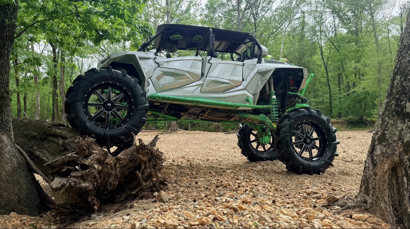 White and green custom side-by-side with oversized off-road tires on a wooded trail